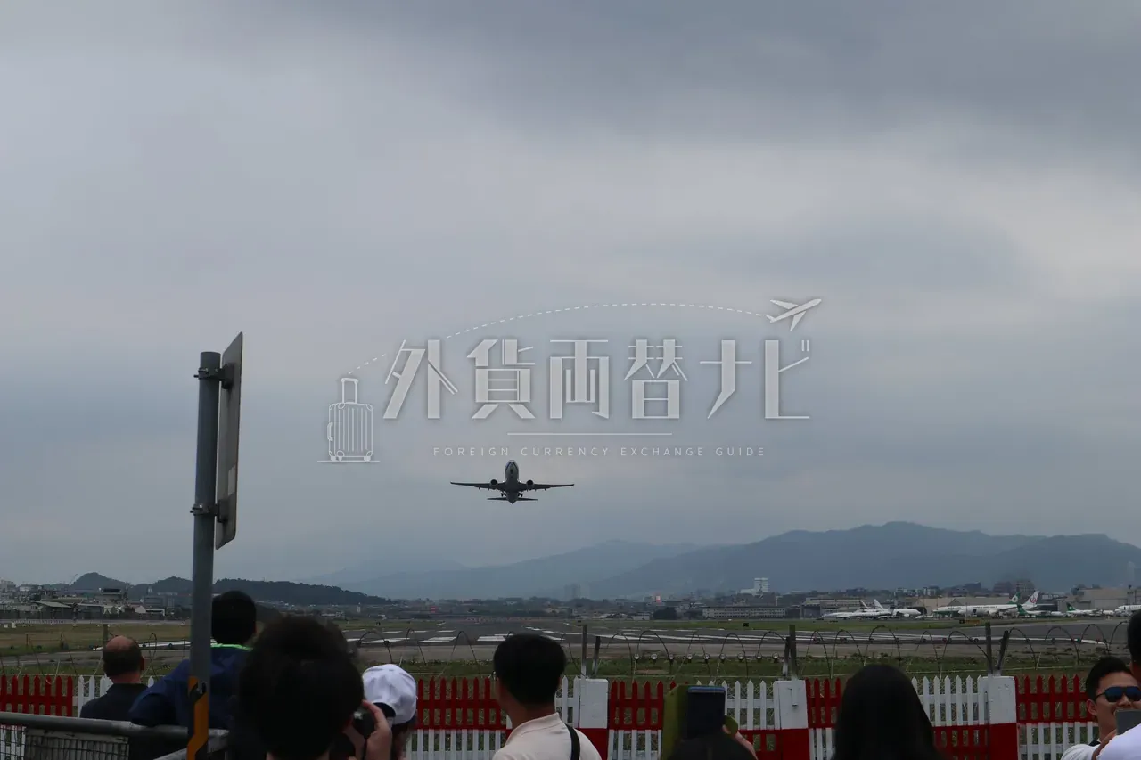 松山空港の離陸滑走路と背景の山並み。赤白柵越しに飛行機を見る人々
